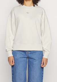 Felpa beige chiaro con scollatura tonda e maniche lunghe, leggermente arricciate. Presenta un piccolo logo ricamato sul petto. Abbinata a jeans blu.