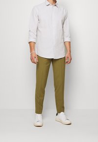 Camicia grigia chiaro a maniche lunghe con bottoni, abbinata a pantaloni verde oliva e sneaker bianche. Caratteristiche includono maniche arrotolabili e design slim fit.