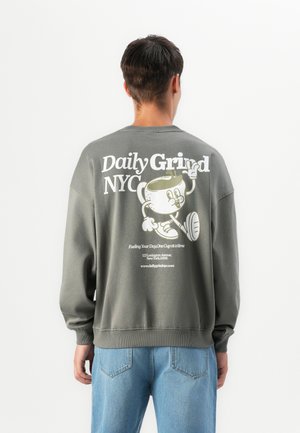 Person mit grauem Sweatshirt, auf dem eine Cartoon-Kaffeetasse und der Text "Daily Grind NYC" auf dem Rücken abgebildet sind, kombiniert mit blauen Jeans, mit dem Rücken zur Kamera gerichtet.
