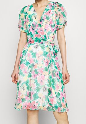 Mujer con un vestido cruzado floral con mangas cortas abullonadas hasta la rodilla, con colores verde, rosa, amarillo y blanco, atado a la cintura.