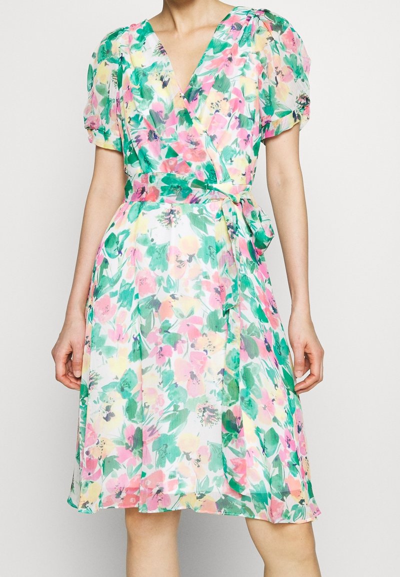 Femme portant une robe portefeuille fleurie à manches courtes bouffantes, longueur genou, dans des couleurs vert, rose, jaune et blanc, nouée à la taille.