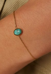 Gouden armband met een ovale turkooise steen met een gebarsten oppervlak, gezet in een gestructureerde zetting, bevestigd aan een fijne ketting.