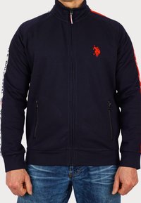 Marintröja med dragkedja i marinblått och röda detaljer, med sidofickor, ribbade ärmslut och en liten logga på bröstet; tillverkad av ett mjukt tyg.