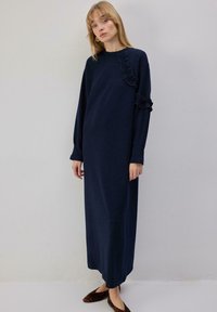 Robe bleu marine à manches longues, avec un détail à volants sur l'épaule gauche. Tissu doux, coupe ample, longueur cheville, portée avec des chaussures plates.