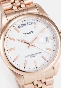 Montre Timex en or rose avec un cadran blanc, affichage jour-date et marqueurs d'heure. Dotée d'un bracelet à maillons et d'une résistance à l'eau jusqu'à 5 ATM.