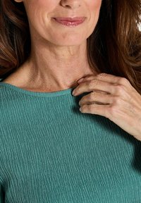 Frau mit langen braunen Haaren, trägt ein türkisfarbenes, strukturiertes Oberteil, berührt mit der rechten Hand ihren Ausschnitt, zeigt natürliche Haut und ein dezentes Lächeln.