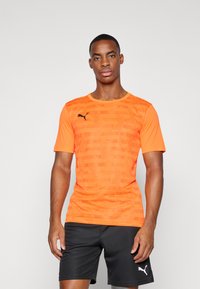 PUMA orange kurzärmliges Sportoberteil mit einem strukturierten Muster, rundem Ausschnitt und schwarzem Logo auf der Brust. Getragen mit schwarzen Shorts.