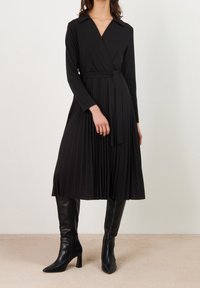 Černé plisované zavinovací šaty s dlouhými rukávy, límcem a páskem stejné látky v pase. Kombinované s černými kozačkami na podpatku po kolena. Hladká textura látky.