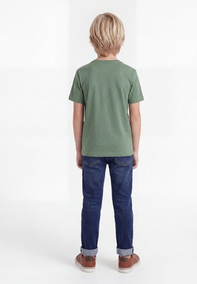 Ξανθό παιδί που στέκεται με την πλάτη, φοράει πουκάμισο ελιάς με κοντά μανίκια, σκουρομπλε τζιν με γυρισμένα μπατζάκια και καφέ αθλητικά παπούτσια σε λευκό φόντο.