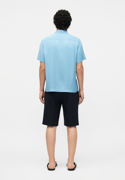 Άνδρας με κατσαρά μαλλιά που φοράει ανοιχτό μπλε κοντομάνικο πουκάμισο, μαύρο σορτς και μαύρα παπούτσια χωρίς κορδόνια, στέκεται με την πλάτη σε λευκό φόντο.