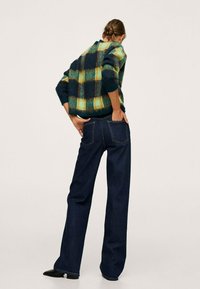 Wollen groene en marineblauwe geruite trui, oversized pasvorm, gecombineerd met donkerblauwe wijde jeans, zichtbare achterzakstiksels, zwarte puntige schoenen.