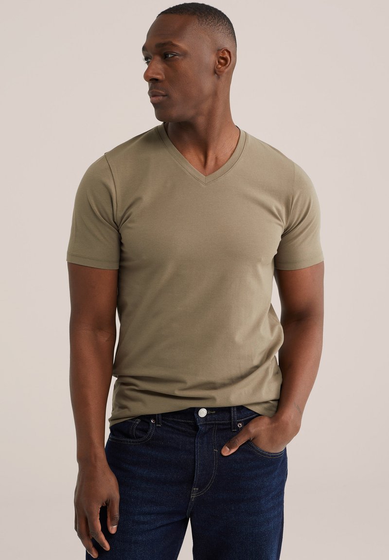 Hombre con camiseta de cuello en V de color verde oliva y vaqueros azul oscuro, mirando hacia su izquierda con una mano en el bolsillo.