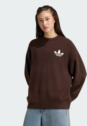 Jeune femme aux longs cheveux foncés portant un pull marron avec un logo Adidas blanc et un jean bleu clair, debout devant un fond uni.