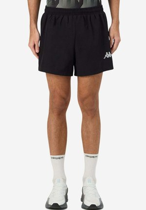 Jambes d'homme portant un short noir Kappa, des chaussettes blanches Kappa mi-mollet et des chaussures de sport blanches, debout devant un fond uni.