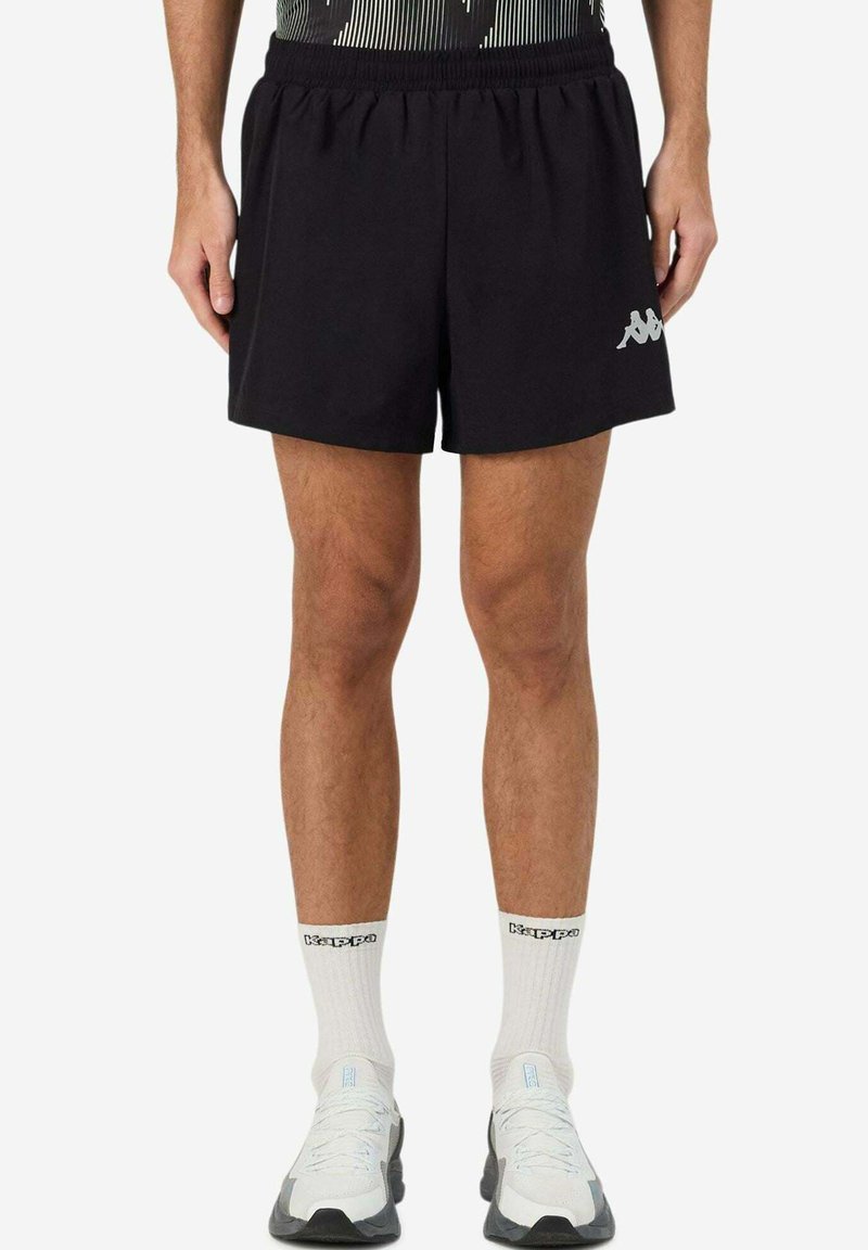 Jambes d'homme portant un short noir Kappa, des chaussettes blanches Kappa mi-mollet et des chaussures de sport blanches, debout devant un fond uni.