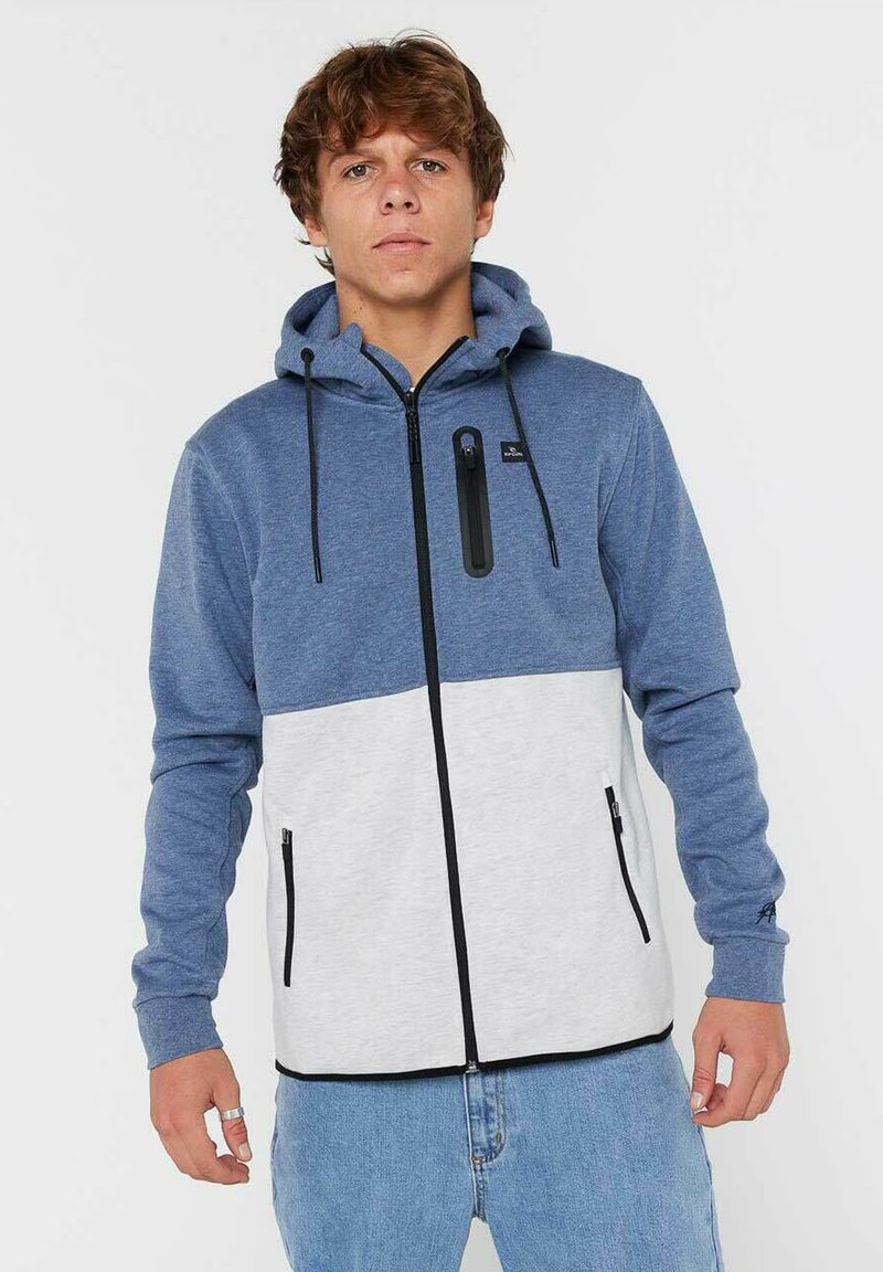 Giovane uomo che indossa una felpa con zip blu e grigia chiara, con dettagli in zip nera, e jeans blu chiaro, in piedi contro uno sfondo semplice.