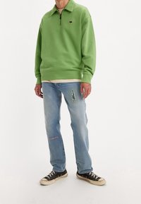 Felpa verde chiaro con cerniera e collo, abbinata a jeans blu strappati e sneakers nere in tela. Logo del marchio ben visibile sul petto.