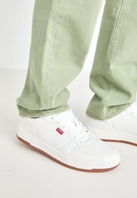 Weiße Sneakers mit perforierten Details und einer braunen Gummisohle, kombiniert mit leichten grünen Baumwollhosen.