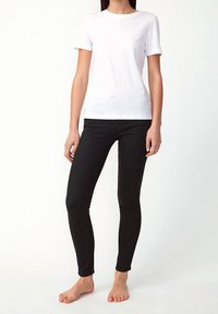 Bílá bavlněná tričko s krátkými rukávy, spárované s černými úzkými džíny. Modelka stojí naboso, ruce má podél těla v neutrální pozici.