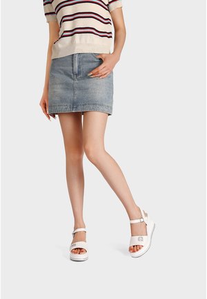 Donna con top a righe maniche corte, minigonna in denim e sandali bianchi con cinturino alla caviglia e tacchi a zeppa bassi, in piedi con una gamba incrociata.