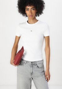 T-shirt bianco a coste con un piccolo logo, abbinato a pantaloni in denim grigi chiari a vita alta. Tiene una borsa a tracolla rossa. Capelli ricci.
