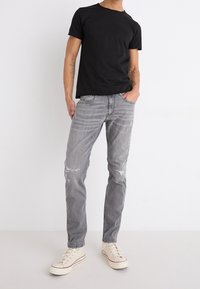 Camiseta negra de manga corta, vaqueros de mezclilla grises desgastados, zapatillas de tono claro con cordones blancos, en pose de pie contra un fondo liso.