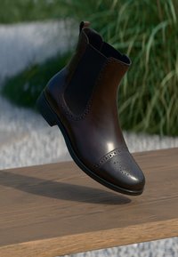 Ruskeat nahkaiset chelsea-saappaat, joissa on rei'itetty kärki ja brogue-yksityiskohtia. Varusteena joustavat sivupaneelit ja matala korko. Suora pinta.