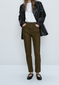 Braune, zulaufende Hosen aus Stoff, kombiniert mit einem weißen T-Shirt und einer schwarzen übergroßen Lederjacke, abgerundet mit schwarzen Ballerinas.