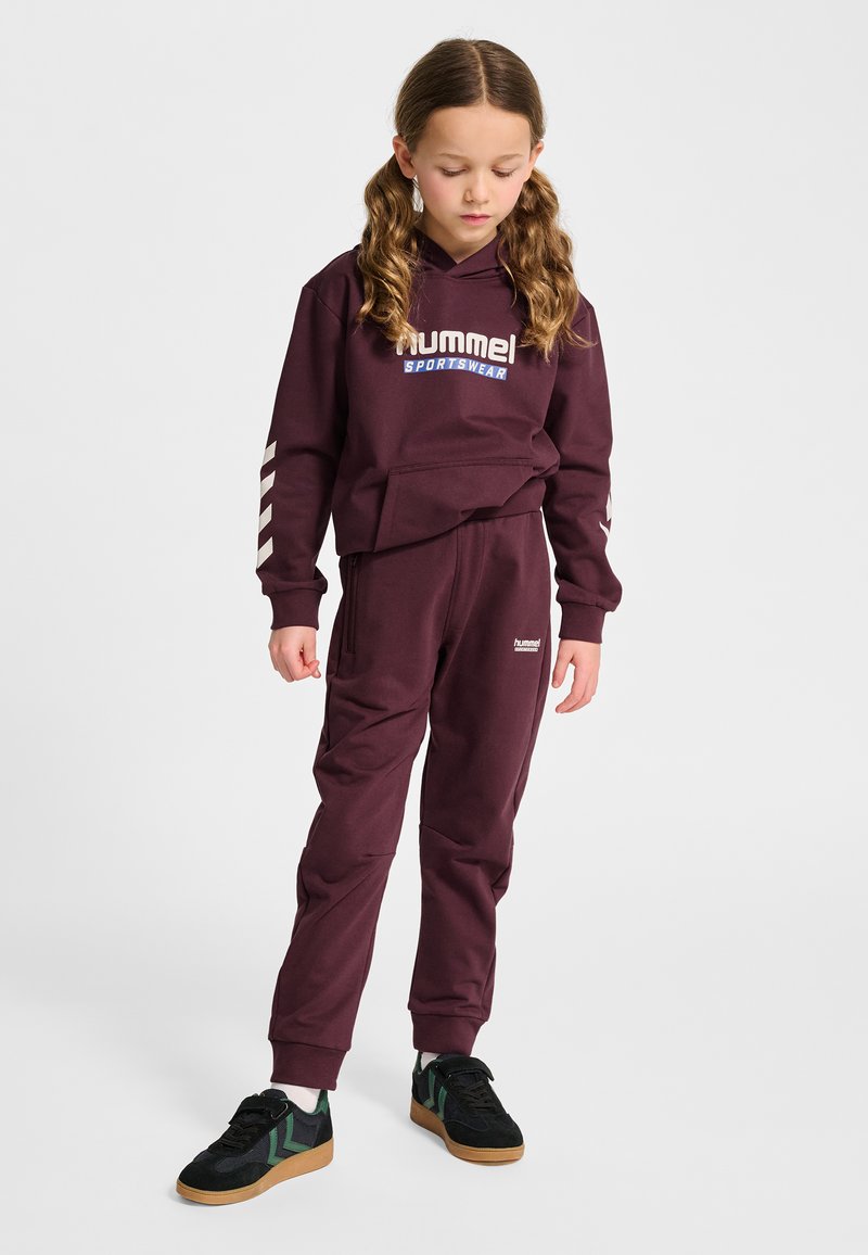 Jong meisje met bruin haar in vlechtjes, gekleed in een maroon hummel sportoutfit en zwarte sneakers met groene details, kijkt naar beneden.