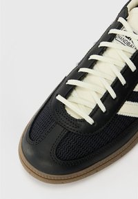 Mornarsko modni športni copat z zgornjim delom iz usnja in tkanine, belimi vezalkami, gumijastim podplatom ter črtastimi poudarki na strani. Klasična atletska zasnova.