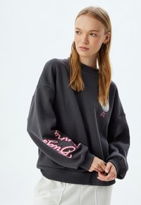 Sudadera gris oscuro con costuras de hombros caídos, puños de canalé y un gráfico colorido en la manga, presentando un escote redondo y una textura suave.