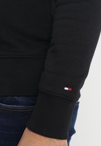 Sudadera negra con puños de canalé, que presenta un pequeño logo tricolor en la manga. Tela texturizada y diseño clásico de cuello redondo.