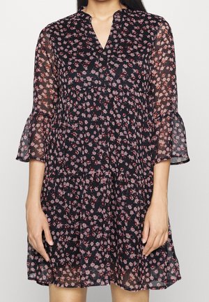 Vrouw draagt een zwarte jurk met bloemenprint in roze en rood, een V-hals en doorschijnende klokmouwen van drie kwart lengte.