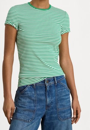 Person trägt ein grün-weiß horizontal gestreiftes kurzärmeliges Shirt, hineingesteckt in hoch taillierte blaue Jeans mit aufgesetzten vorderen Taschen.