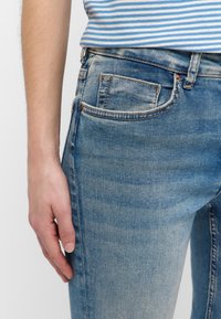 Jeans de mezclilla azul claro con un ajuste slim, que cuentan con un pequeño bolsillo frontal, un desvanecimiento sutil y costuras visibles a lo largo de las costuras.