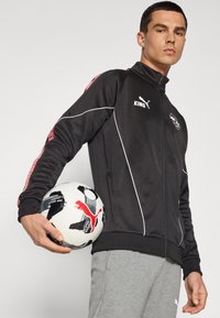 Svart zip-up sportjacka med vita accentueringsdetaljer och röd mönstrade ärmar, bärs över grå byxor, hålls med en svart och vit fotboll.