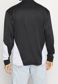 Veste de sport noire avec des panneaux latéraux blancs, manches longues et col. Texture lisse et coupe décontractée. Aucune marque ou motif visible.