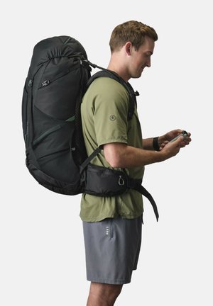 Black, durable backpack with multiple compartments and mesh sides, worn on the back. User in olive shirt and gray shorts, holding a small object.