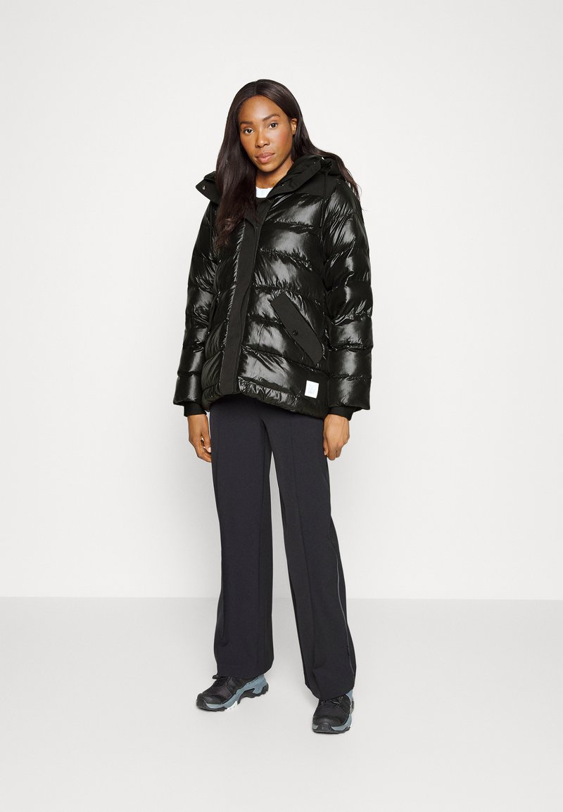 Gepolsterte schwarze Jacke mit glänzender Oberfläche, Reißverschluss, großen Taschen, abnehmbarer Kapuze, getragen über schwarzen Hosen und Schuhen.