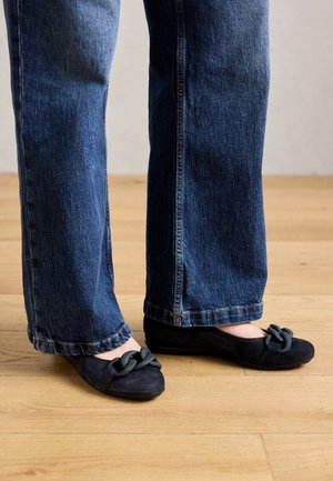 Suede black flats with a knotted detail on the toe, worn with blue wide-leg jeans. Features a smooth texture and a low-profile design.