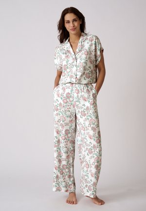 Vrouw staat op blote voeten, draagt een losse witte pyjamaset met bloemenprint, korte mouwen en wijde broekspijpen, handen in de zakken.