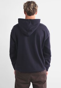 Sweat-shirt bleu marine avec capuche, doté d'une coupe décontractée et de poignets côtelés. Le matériau semble doux et texturé, avec un design simple.