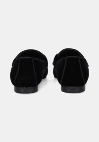 Par de mocasines de terciopelo negro sin cordones con tacones bajos, vistos desde atrás sobre un fondo blanco plano.