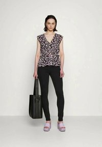 Top preto e lavanda com padrão floral e mangas curtas, combinado com calças pretas justas e sapatos de plataforma lilás claros. Segura uma bolsa tote preta.