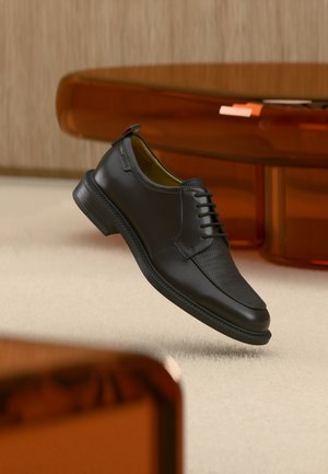 Scarpa elegante da uomo in pelle nera con stringhe e parte anteriore texturizzata, sospesa sopra un tappeto beige in una stanza moderna con mobili dalle tonalità ambra.