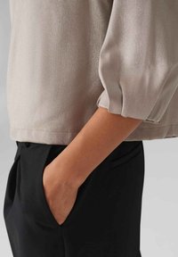 Seidiger, hellbeiger Oberteil mit weiten, gerafften Ärmeln, kombiniert mit schwarzen Hosen mit schräge Taschen. Glatte Textur, schlankes Design.