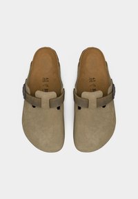 Ζευγάρι μπεζ σουέντ παντόφλες με ρυθμιζόμενους καφέ δερμάτινους ιμάντες και πάτους από φελλό, εμφανιζόμενες από ψηλά σε απλό φόντο.