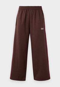 Hnědé sportovní kalhoty s širokým střihem, zdobené burgundským pruhy po stranách a elastickým pasem; zahrnují branding "Kakiko Society".