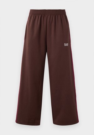 Brune sportbukser med bred bendesign, der har en bordeaux sidelinje og elastisk talje; inkluderer "Kakiko Society" branding.