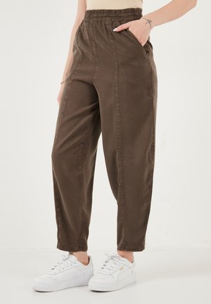 Brązowe spodnie z wysokim stanem i elastyczną talią, z bocznymi kieszeniami, noszone z białymi trampkami na platformie i jasną górą, pokazane od talii do stóp.